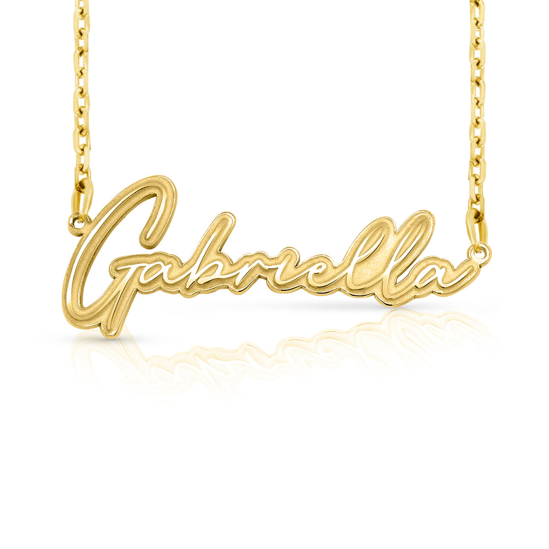 A gold nameplate necklace spelling 'Gabriella' in cursive script, suspended from a delicate gold chain against a white background with a soft reflection below.  This photo can change appearance depending on the name input using the custom name builder