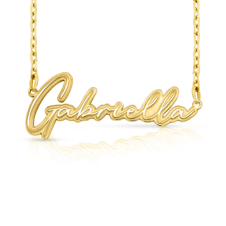 A gold nameplate necklace spelling 'Gabriella' in cursive script, suspended from a delicate gold chain against a white background with a soft reflection below.  This photo can change appearance depending on the name input using the custom name builder