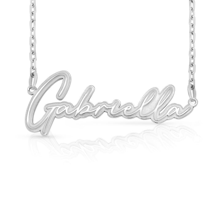 A silver nameplate necklace spelling 'Gabriella' in cursive script, suspended from a delicate silver chain against a white background with a soft reflection below.  This photo can change appearance depending on the name input using the custom name builder