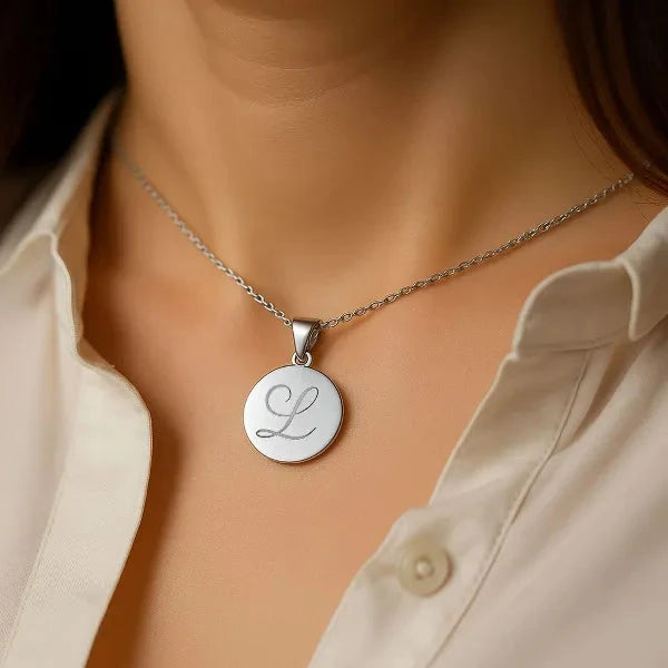 A close-up of a person wearing a silver round pendant necklace with a cursive letter 'L' engraved on it, styled with a cream-colored collared shirt.