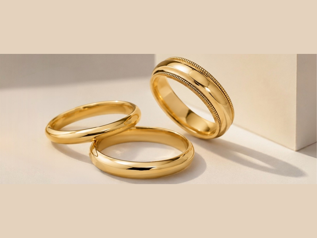 Three polished gold wedding bands arranged on a warm beige surface with a matching block backdrop. Two bands are lying flat and one is standing upright, all showing smooth, reflective surfaces with subtle beaded detailing on the standing band.